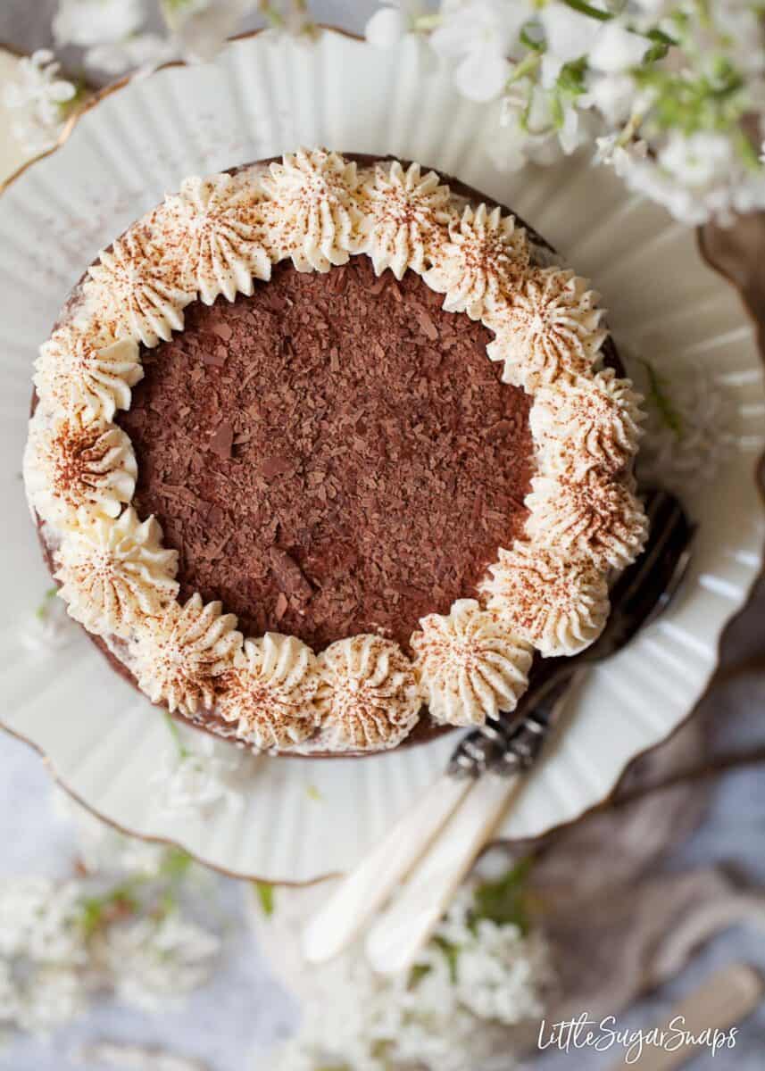 Overhead view of chocolate sponge cake decorated with piped whipped cream, flaked chocolate and cocoa powder. On a gold rimmed fluted plate surrounded by flowers