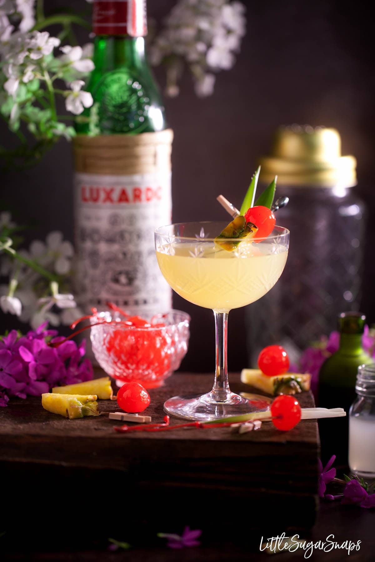 A gin, pineapple and cherry cocktail garnished with fresh pineapple and maraschino cherry. 