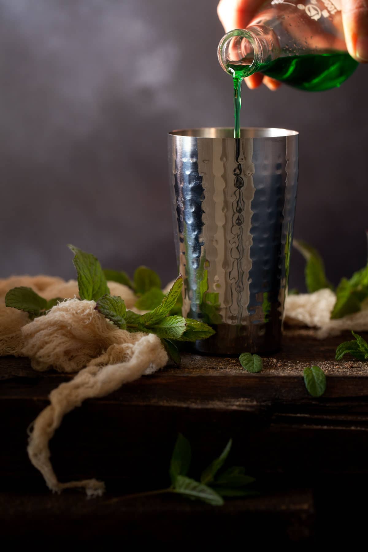 Creme de menthe (green) being poured into a cocktail shaker. There are fresh mint leaves on the wooden table alongside the cocktail shaker