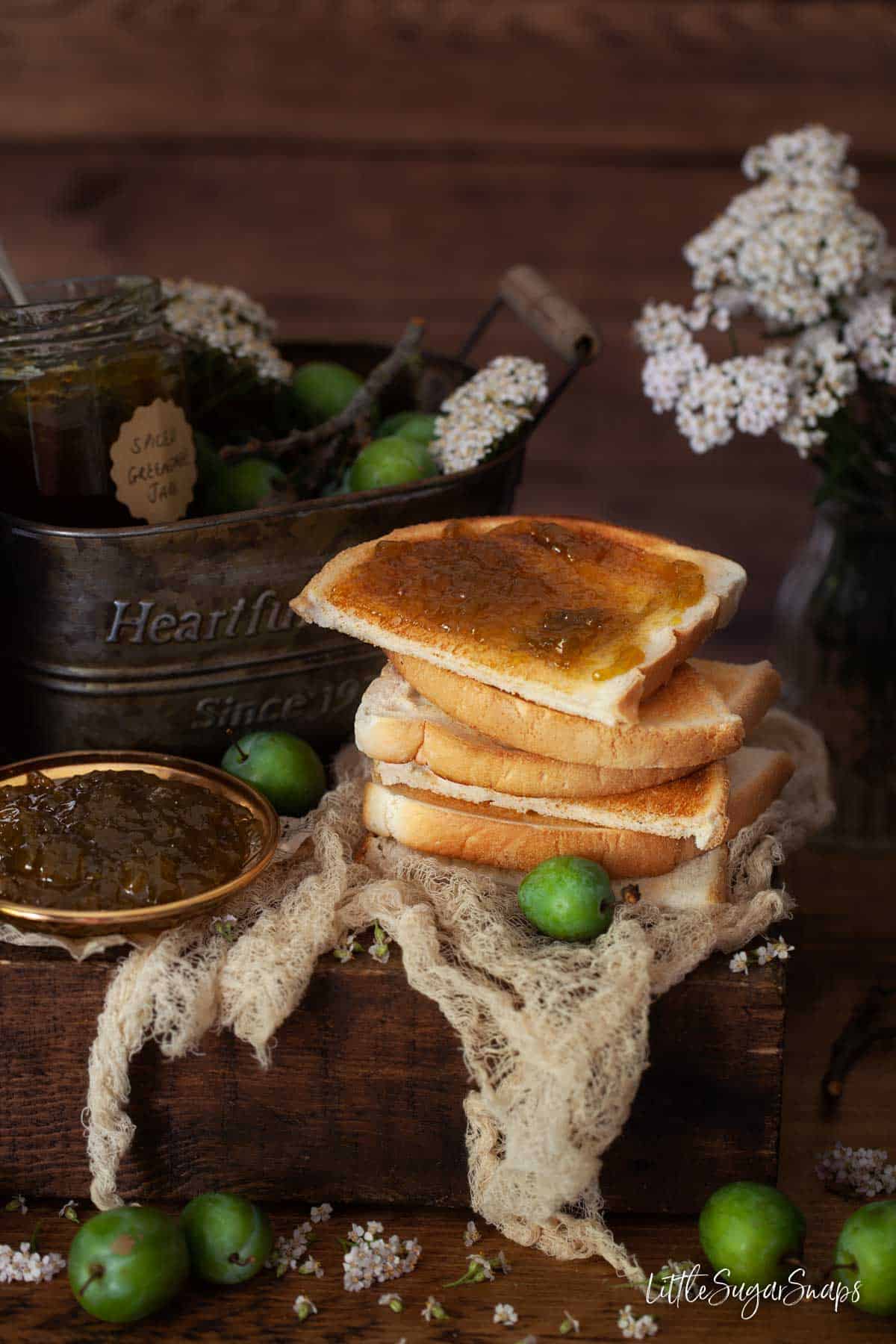 Toast stacked on a wooden board. One slice is spread with greengage jam