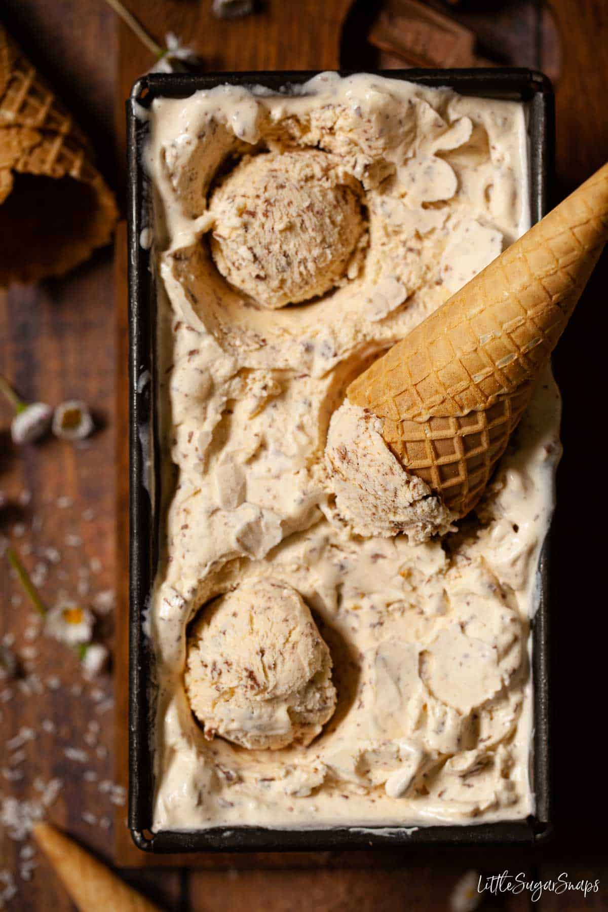 A tub of coconut ice cream with balls scooped out and a waffle cone on top