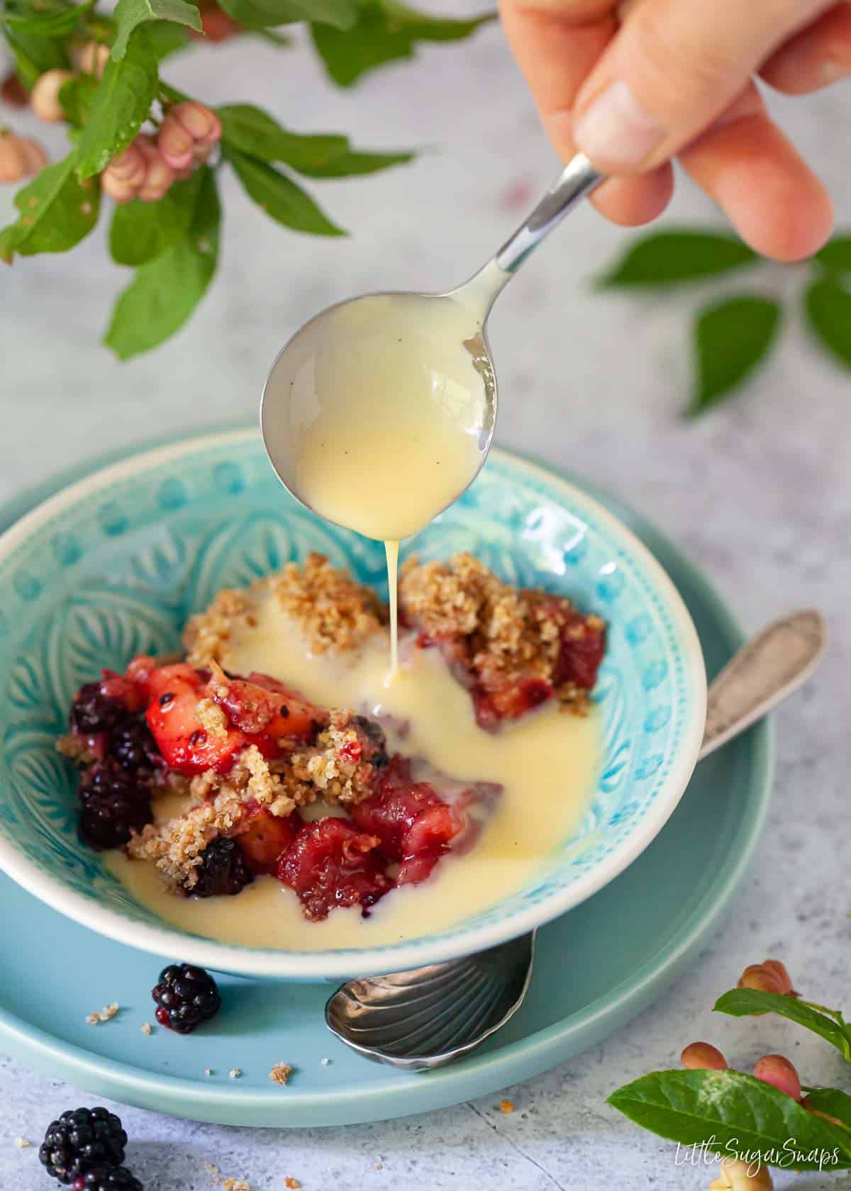 person spooning custard over a serving of apple and blackberry crumble in a bowl