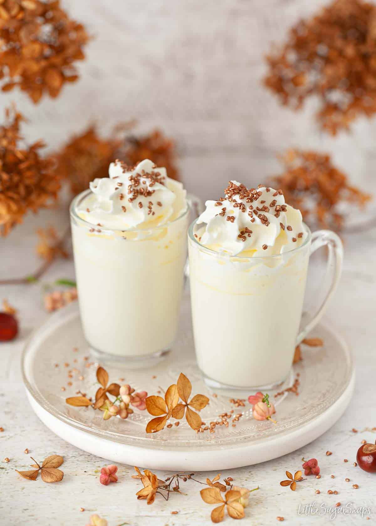 Two glass mugs of white hot chocolate with cream and bronze sprinkles