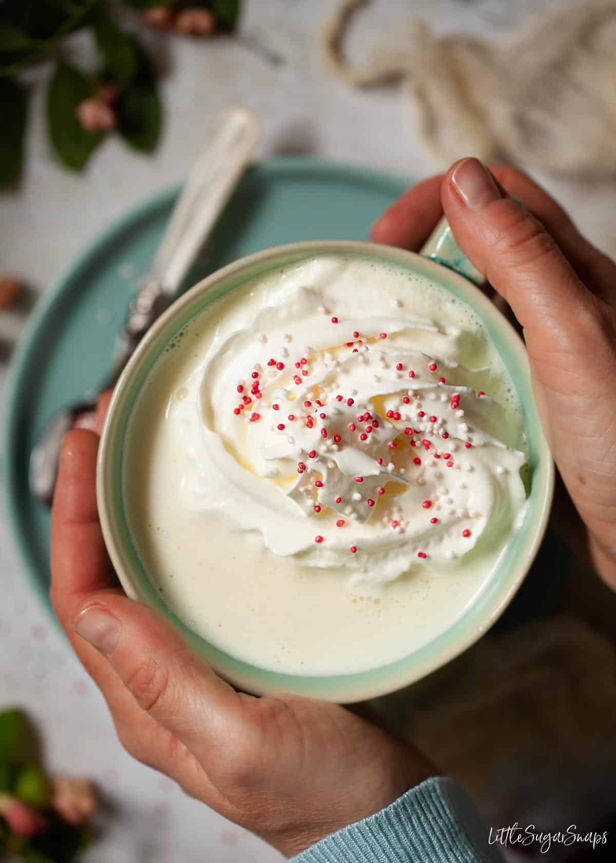 Hands around a mug of white hot chocolate topped with cream and sprinkles