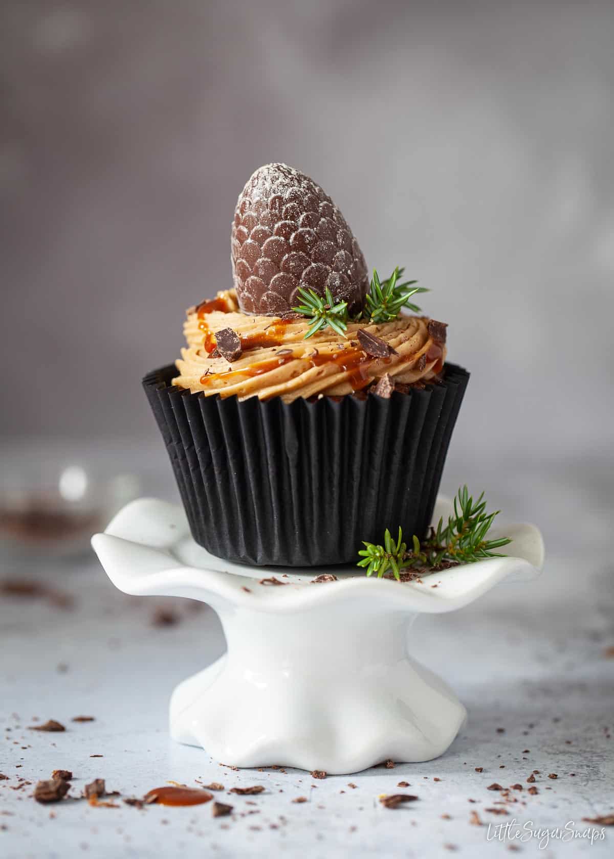 A coffee cupcake on a stand. The cake is topped with buttercream, caramel and a chocolate.