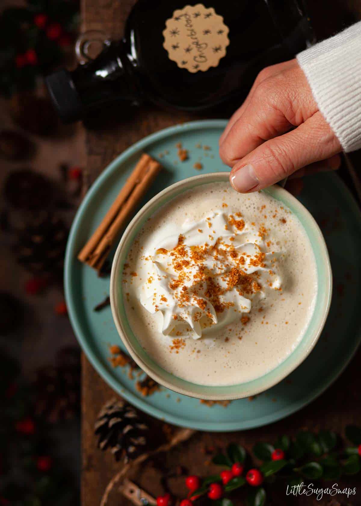 A person about to lift a cup of gingerbread flavoured steamed milk with cream.