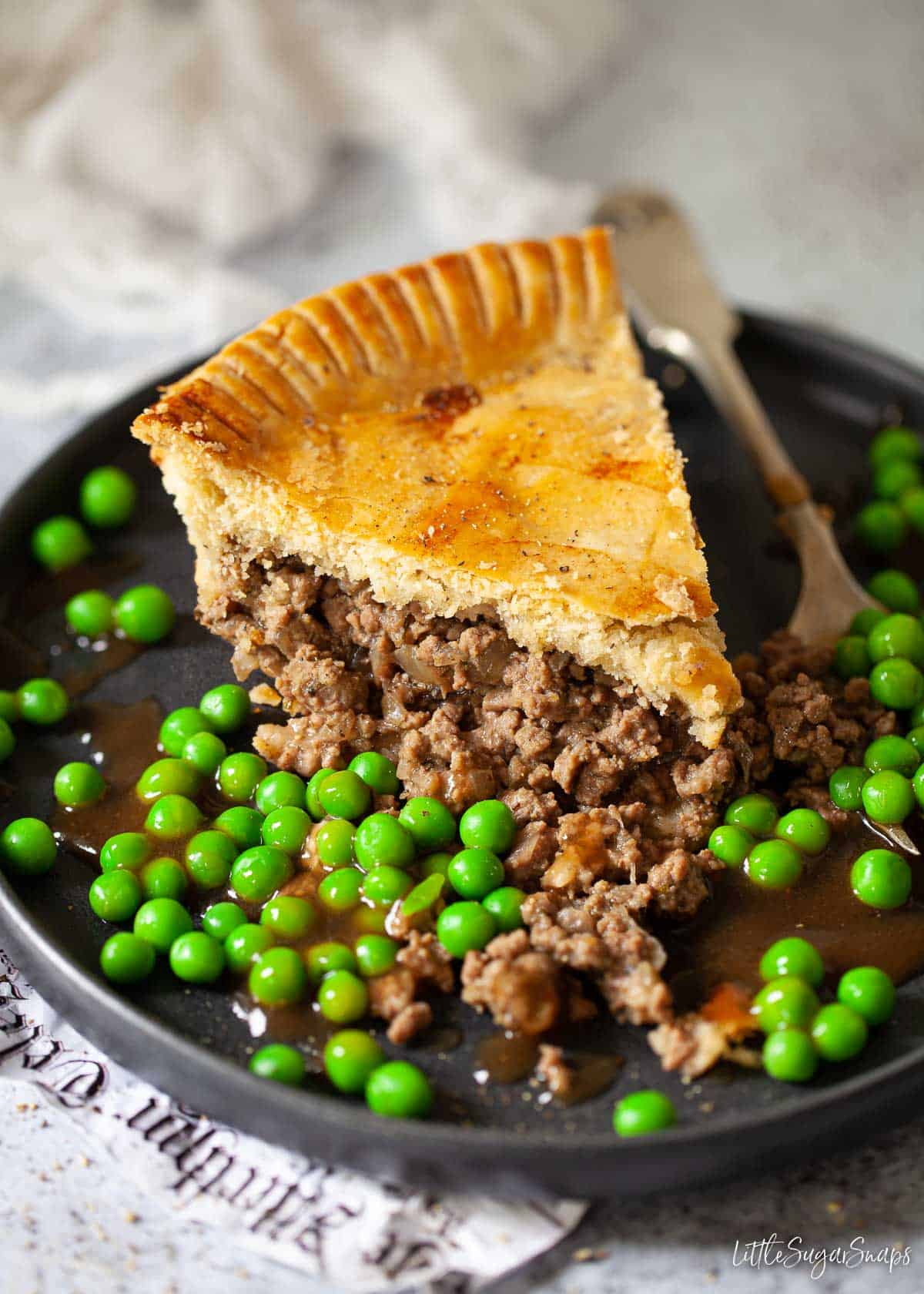 A slice of minced beef and onion pie with peas and gravy.