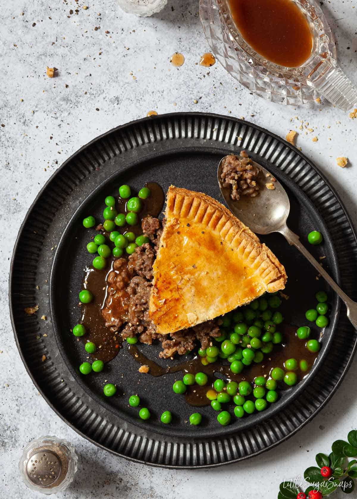 A slice of minced beef and onion pie with peas and gravy on a plate.