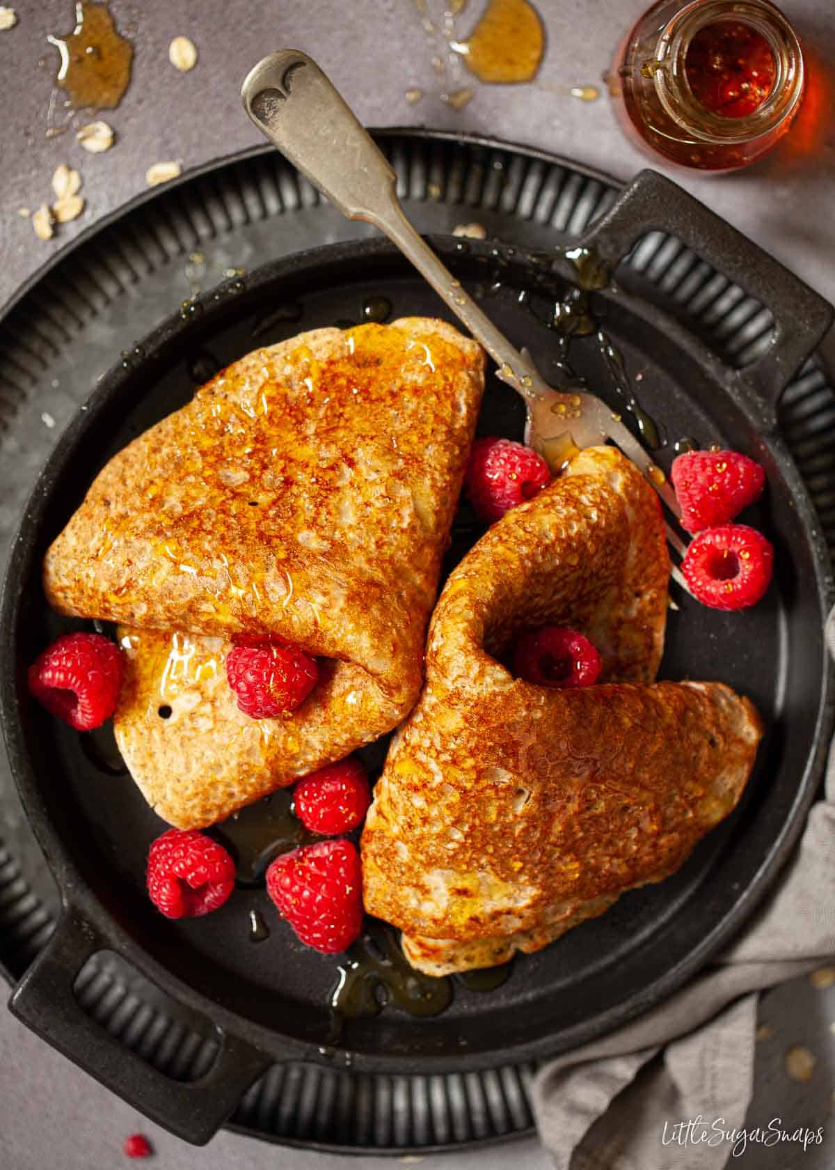 Two North Staffordshire oatcakes on a black plate with golden syrup and raspberries.