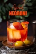 A Negroni cocktail made with Aperol italian Bitters and garnished with orange zest and olives. With text overlay.