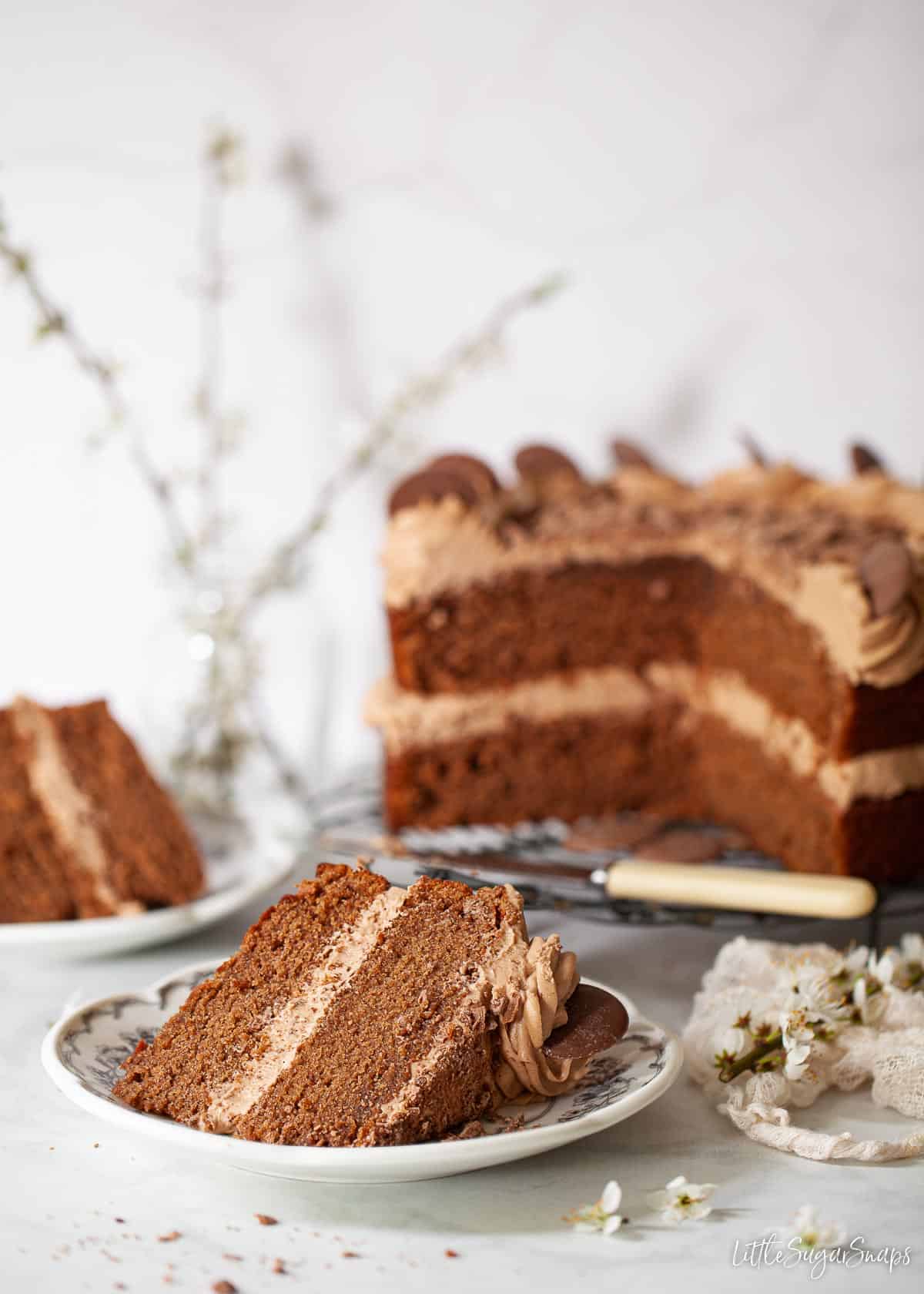 A slice of Dairy Milk Chocolate Cake with the rest of the cake in the background.