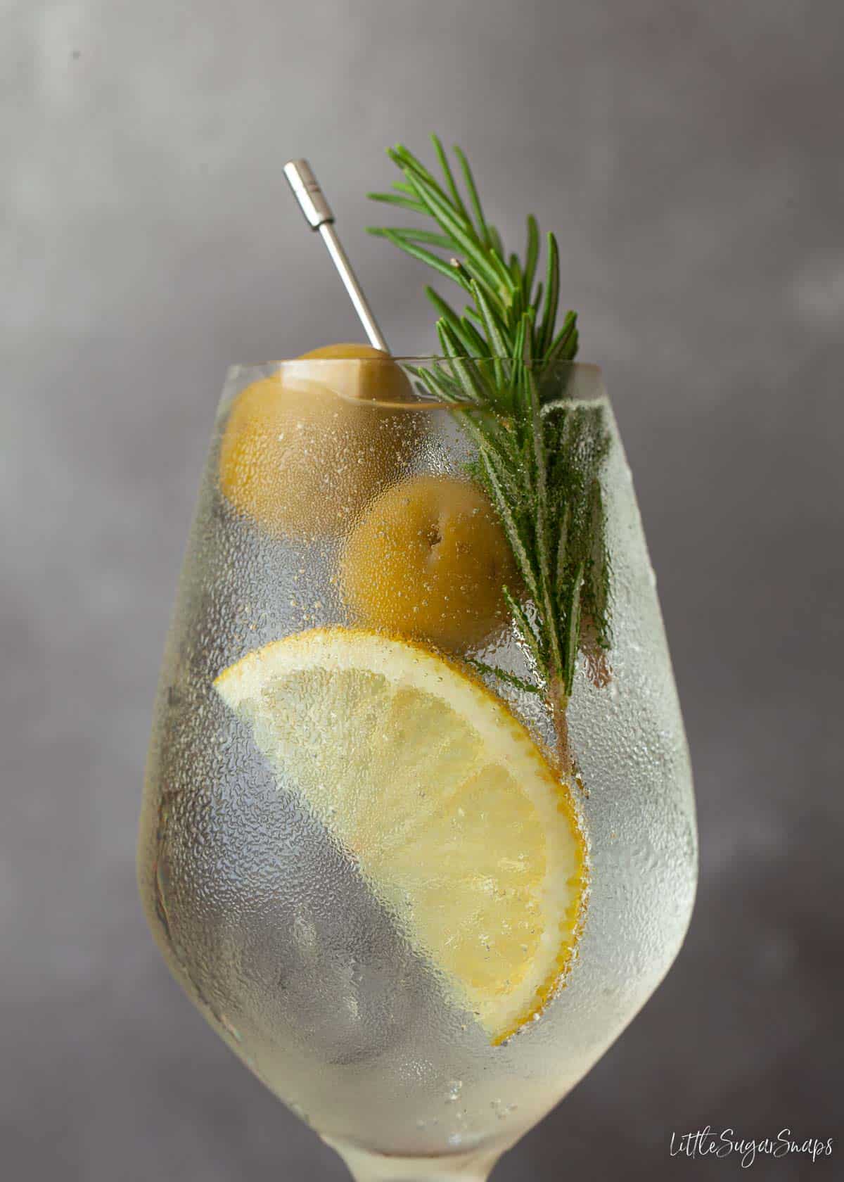 Close up of a white spritz with lemon, rosemary and olive garnish.