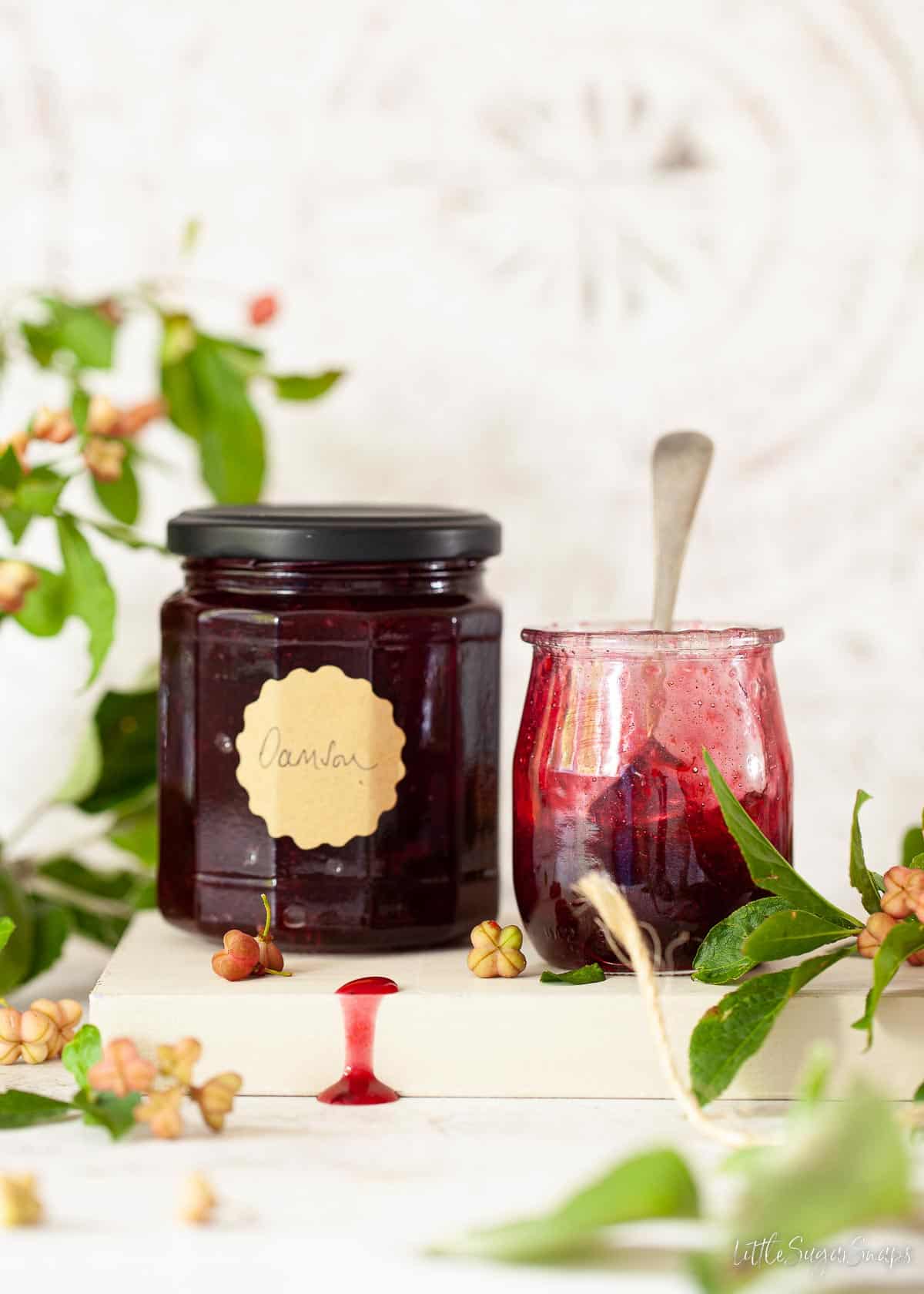 A jar of plum preserve with some it a glass pot alongside.