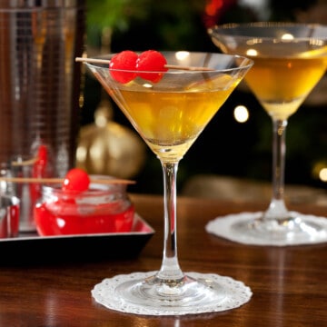 A golden coloured cocktail in a martini glass garnished with Maraschino cherries.