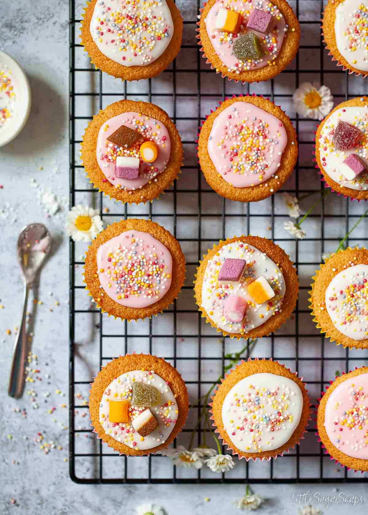 Iced fairy cakes on a wire rack. They are topped with Dolly Mixture sweets and sprinkles.