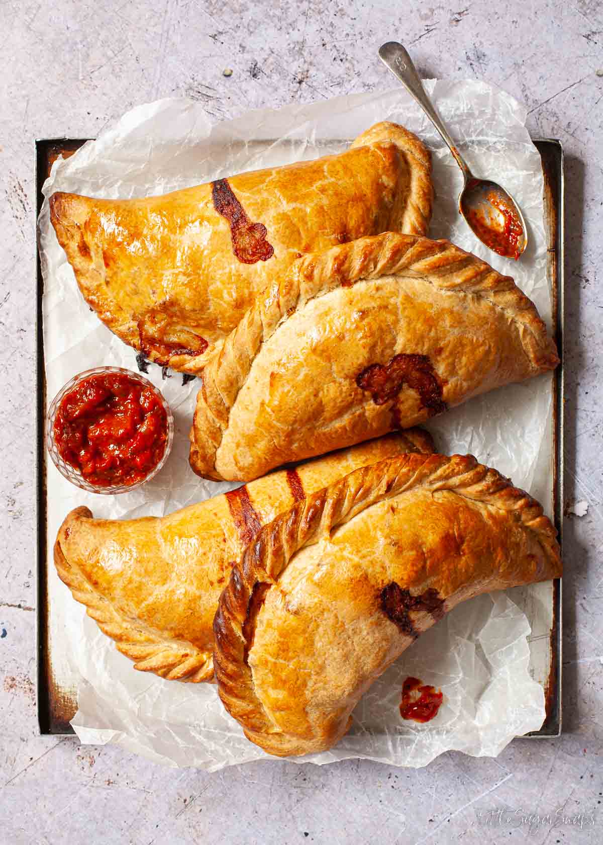 Cheese and onion pasties on a baking tray with a pot of tomato relish.