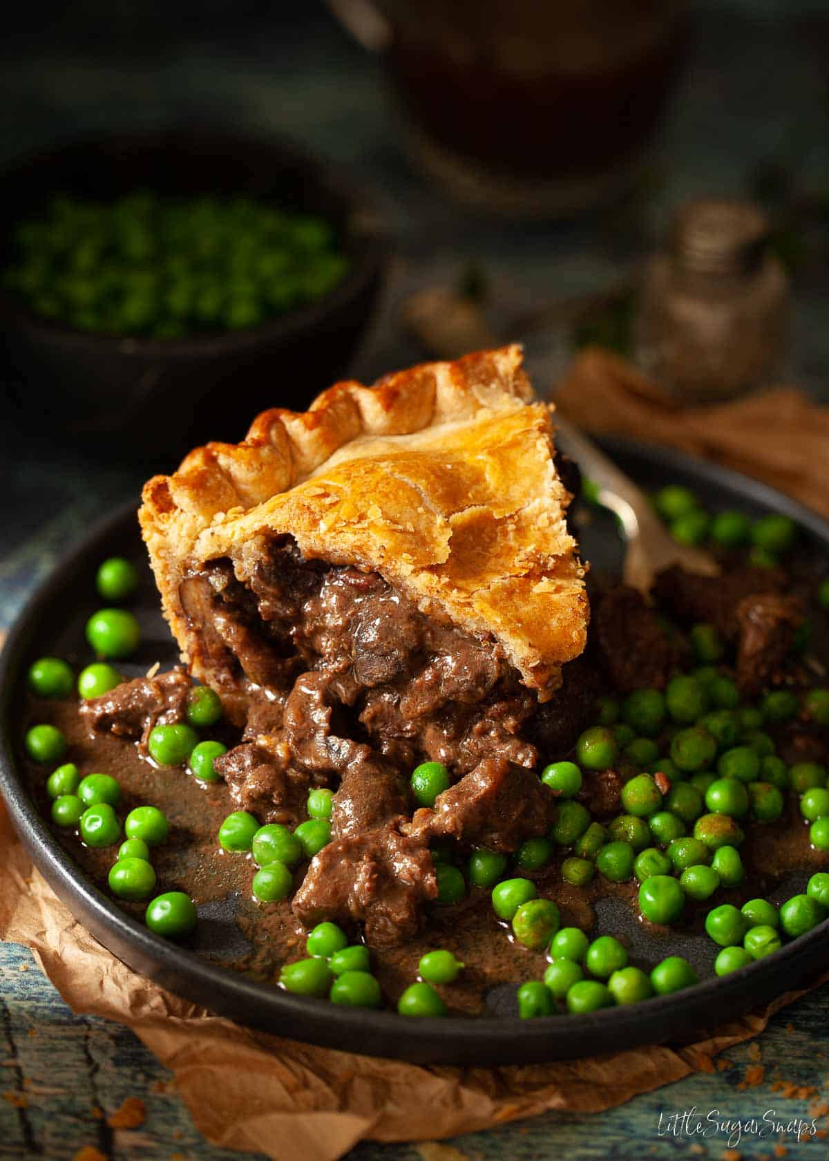 A slice of venison pie on a plate with peas and gravy.