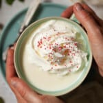 Hands holding a cup of white hot chocolate drink topped with cream and sprinkles.