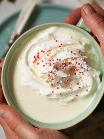 Hands holding a cup of white hot chocolate drink topped with cream and sprinkles.