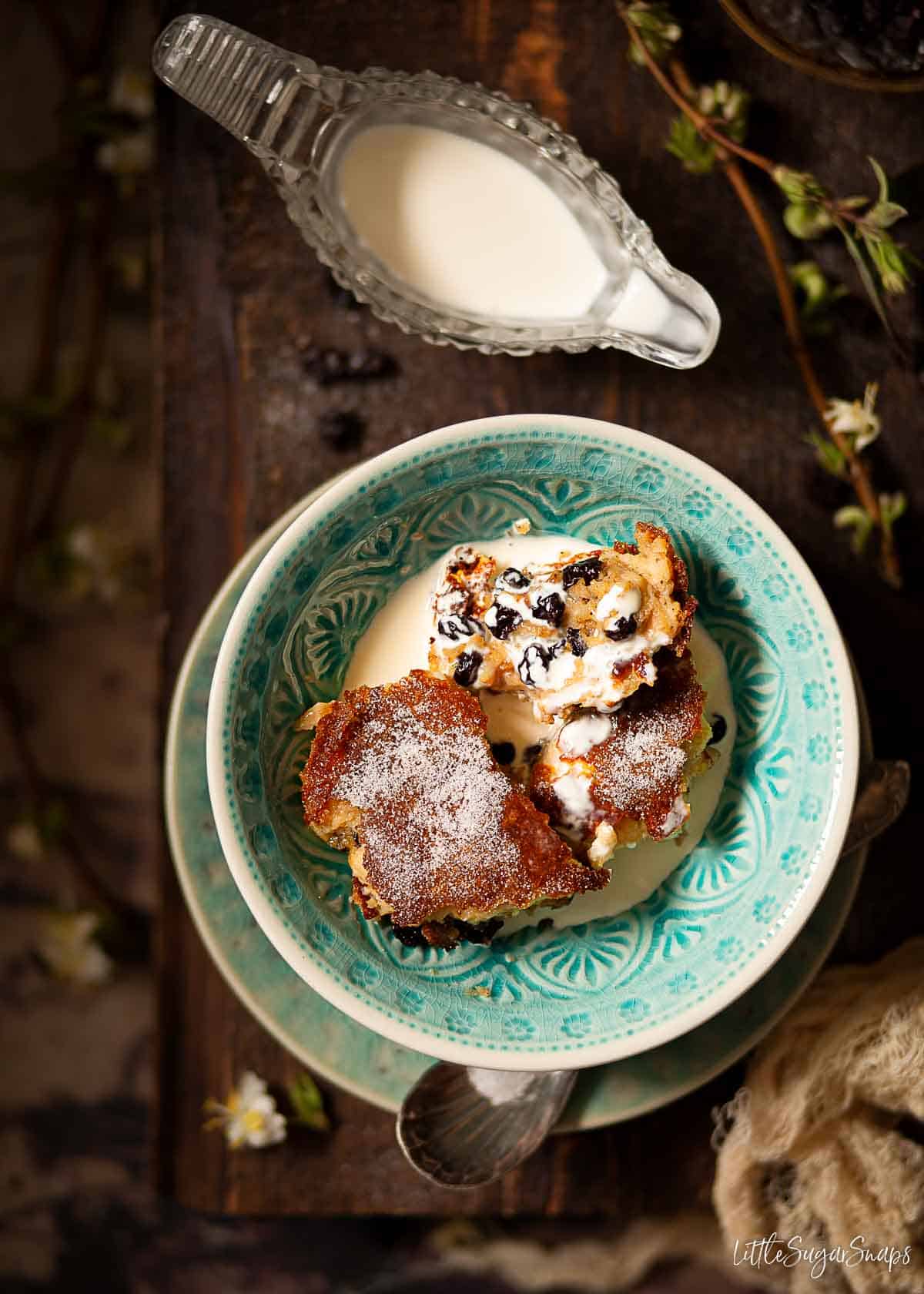 Sweet suet pudding with dried fruit in a blue bowl with runny cream poured over it.