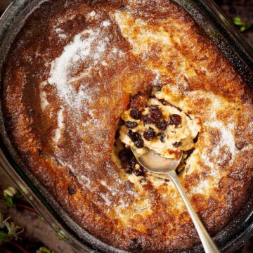 A bowl holding a partially served baked suet pudding with currants. A serving spoon is in the centre.