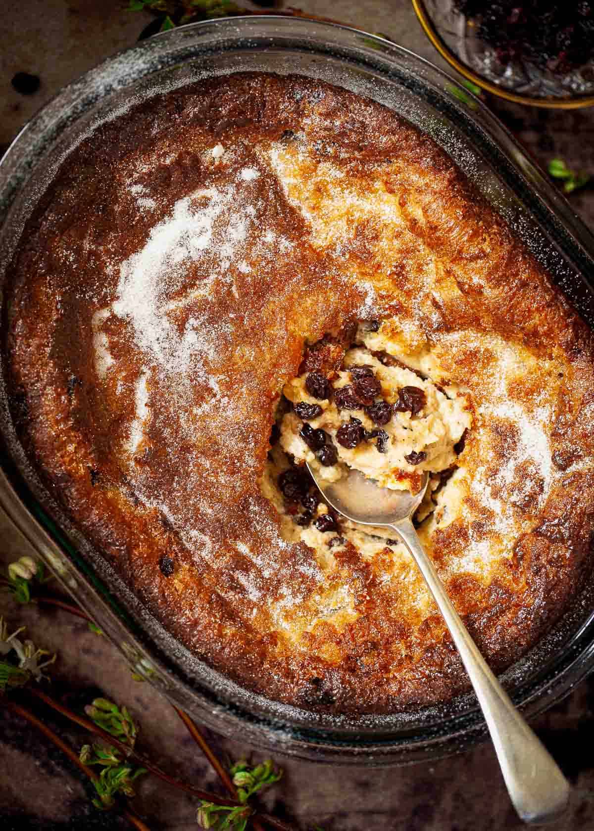 A bowl holding a partially served baked suet pudding with currants. A serving spoon is in the centre.