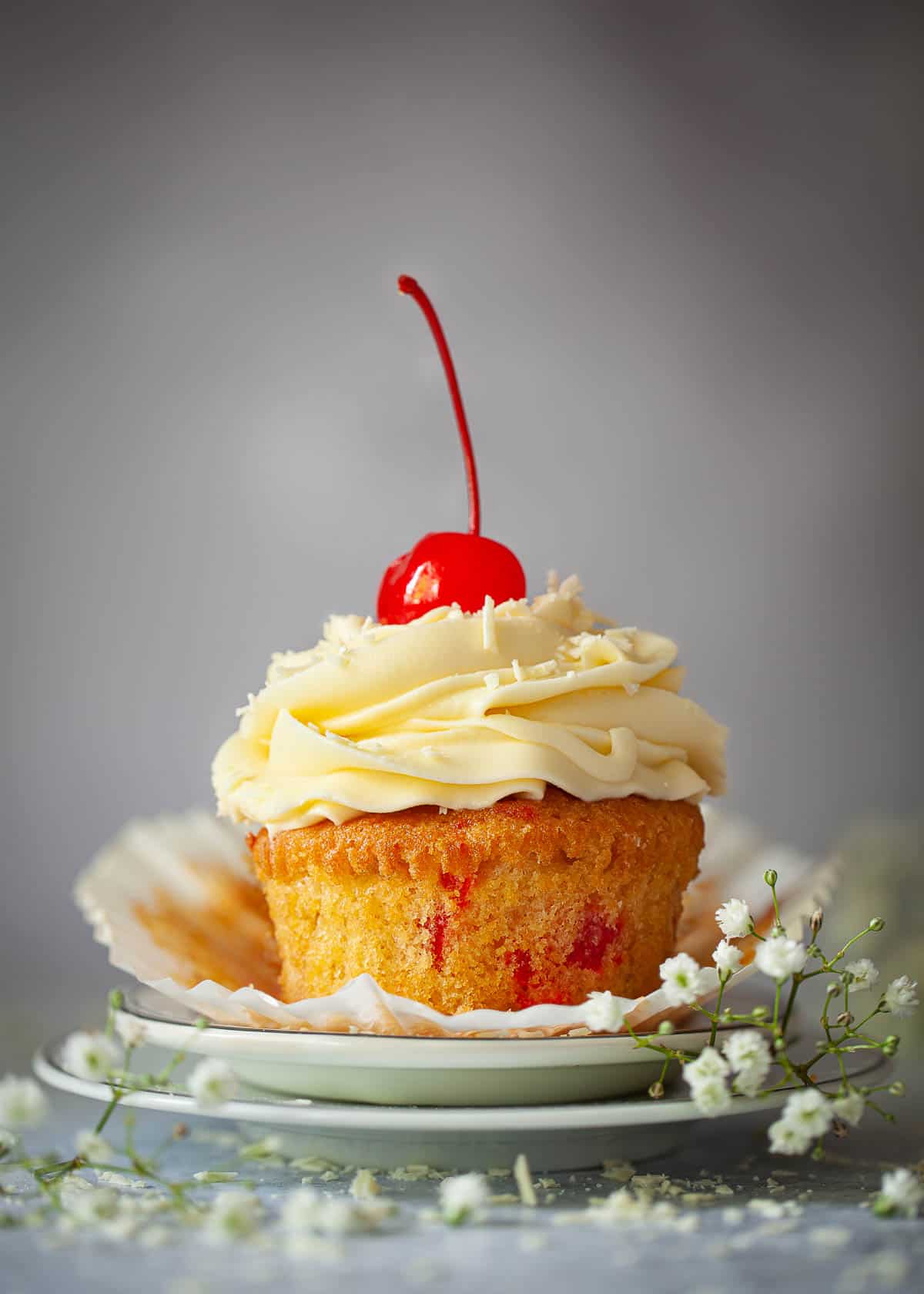 A cupcake with the wrapper pealed away. It has piped buttercream and a maraschino cherry on the top.