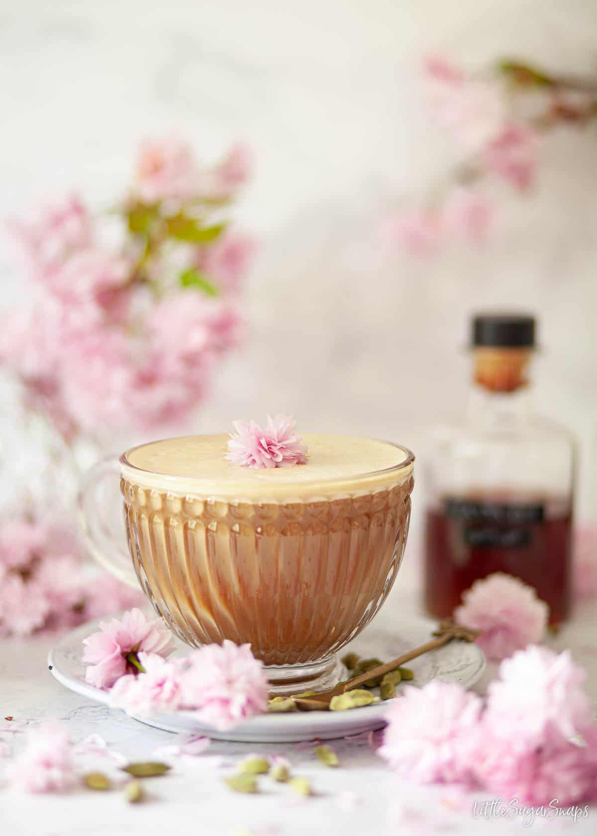 Cardamom latte in a glass cup with a bottle of cardamom syrup and pink blossom.