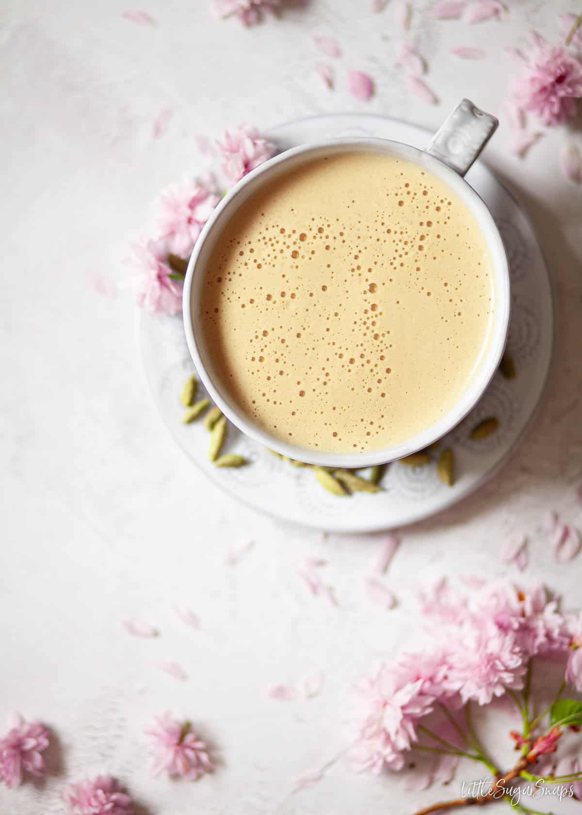 Overhead view of almond milk latte in a wide cup with cardamom pods and pink blossom.