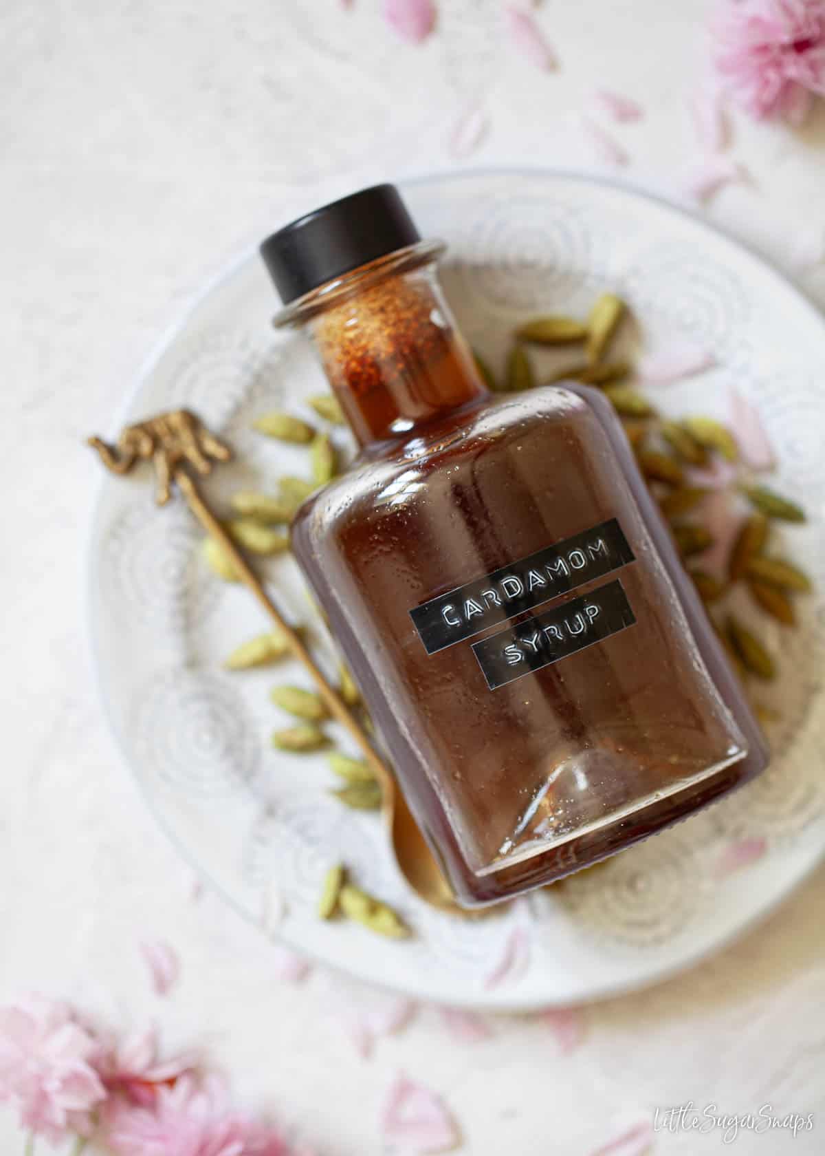 A labelled bottle of cardamom syrup lying on its side on a plate with cardamom pods.