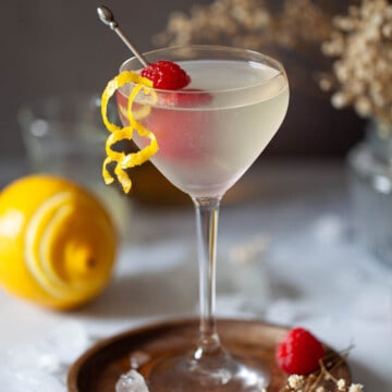 A vodka drink in a Nick and Nora glass garnished with lemon zest and raspberries.
