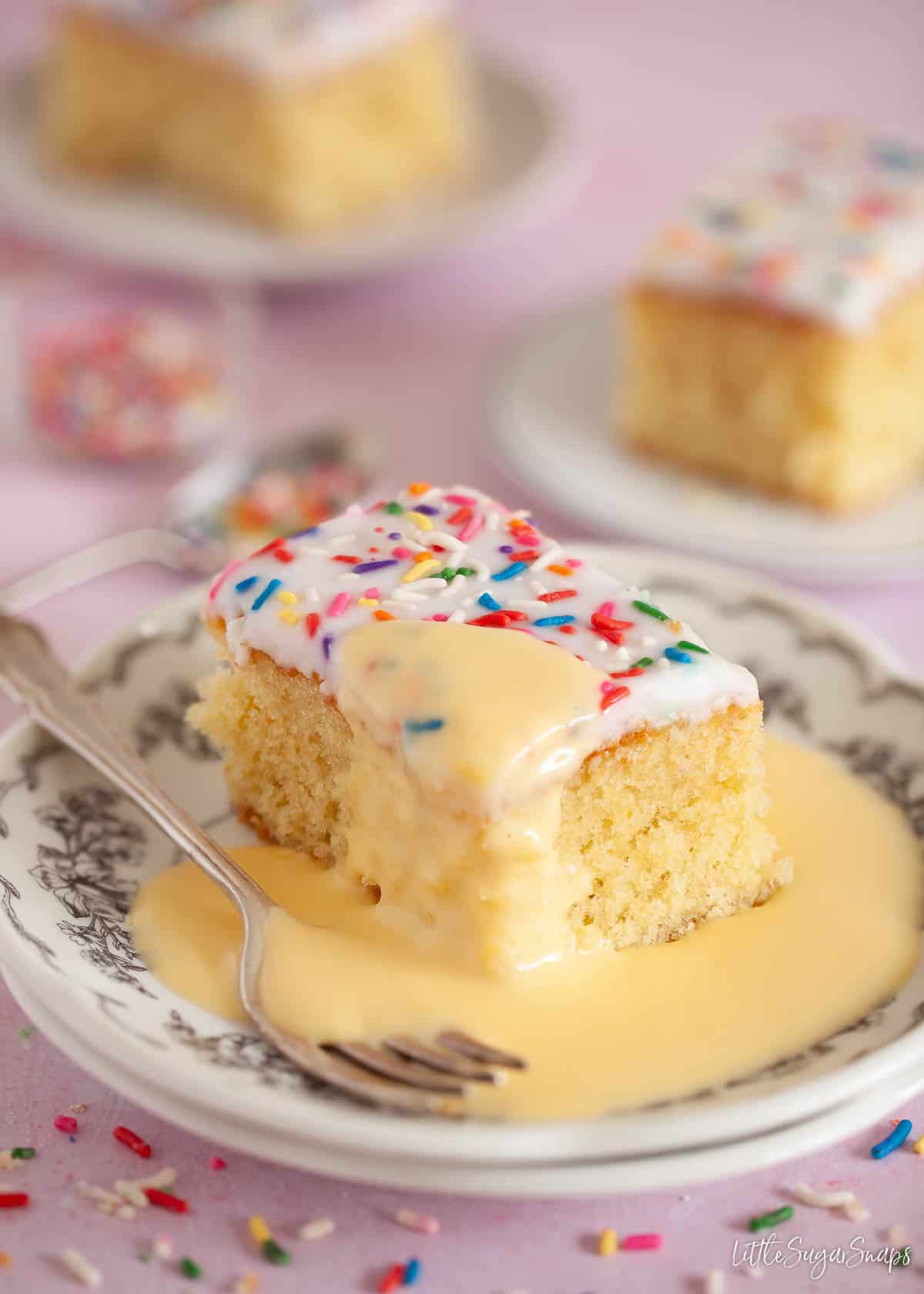 A slice of iced sponge cake topped with sprinkles and served with custard.