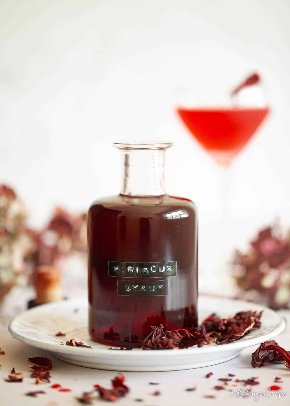 A labelled bottle of hibiscus syrup with a cocktail in the background.