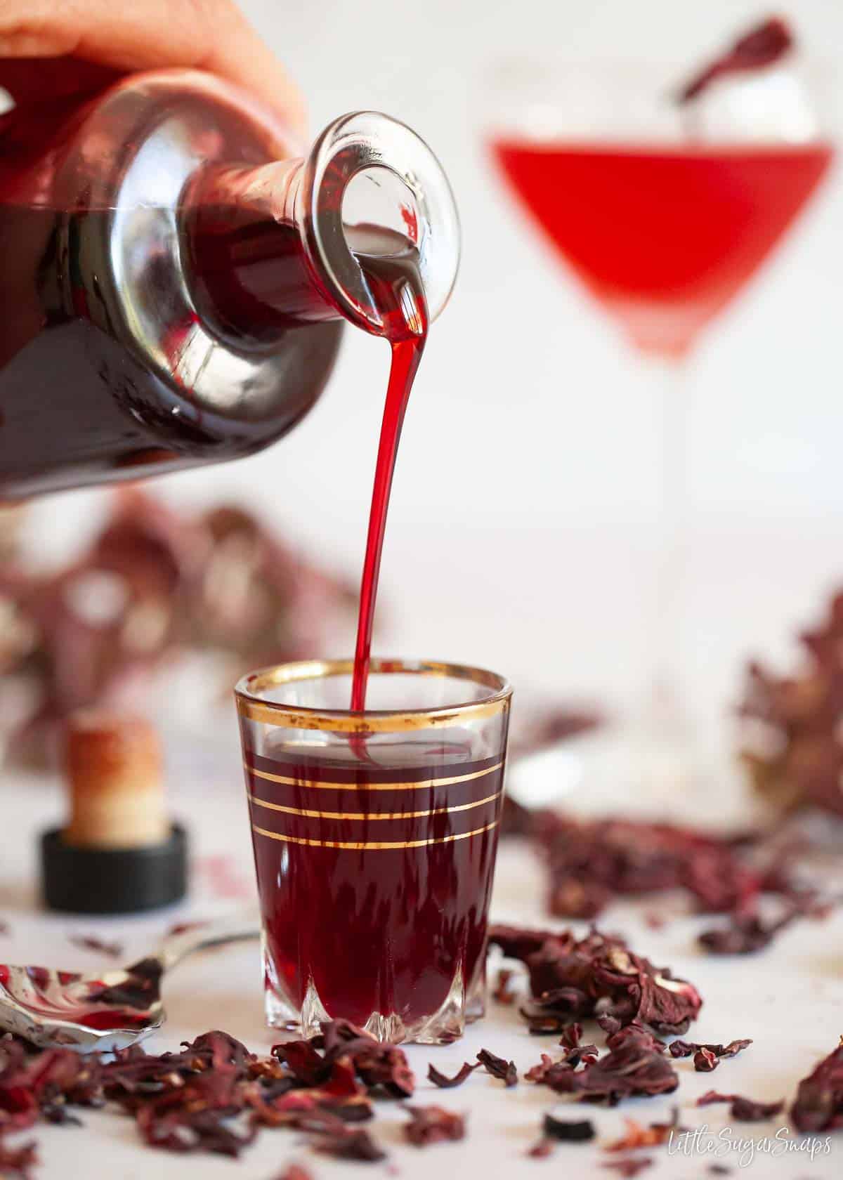 Dark red syrup being poured into a small glass with dried hibiscus flowers on a tabletop.