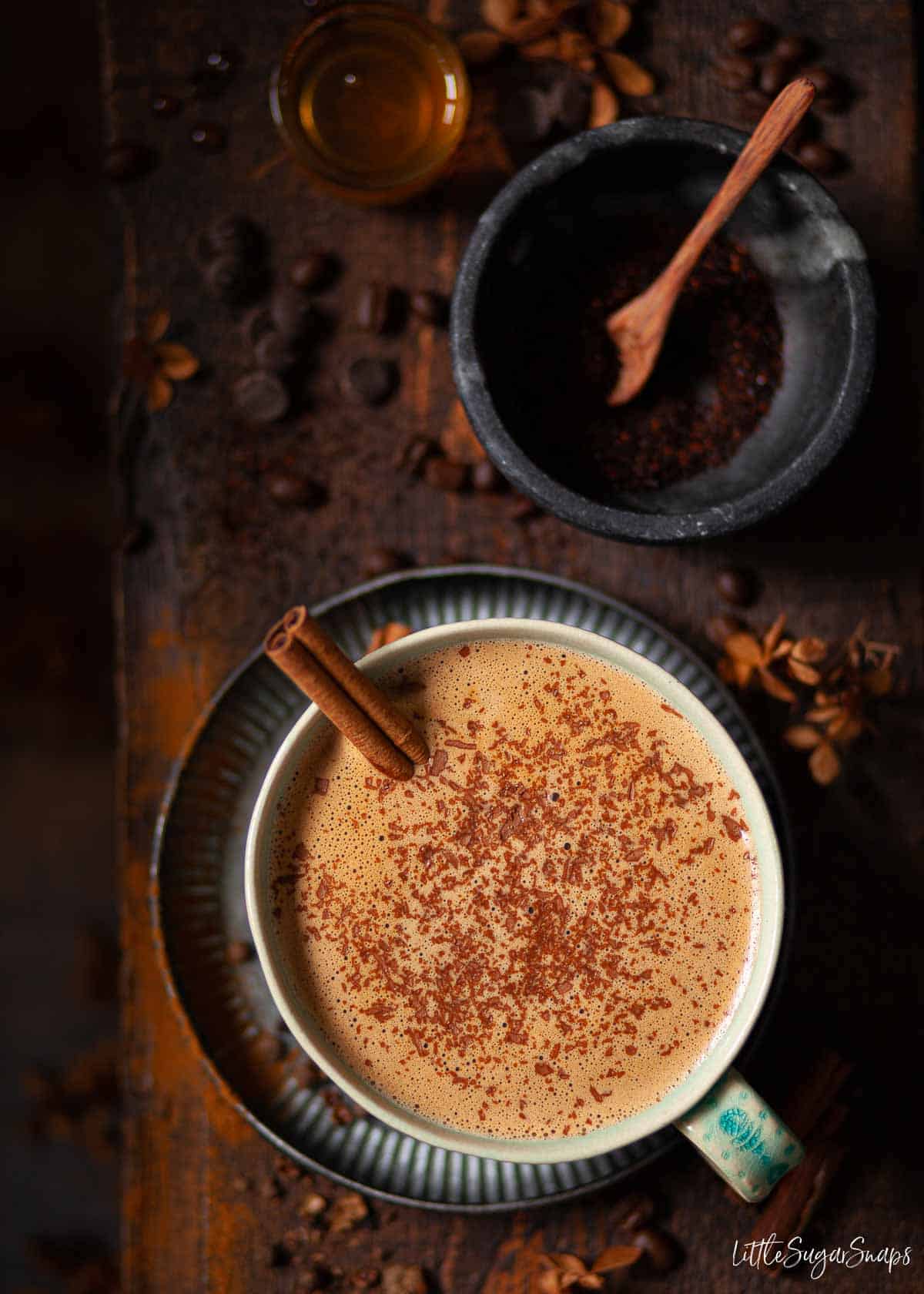 A cup of Mexican mocha topped with grated chocolate and a cinnamon stick with ingredients in shot.