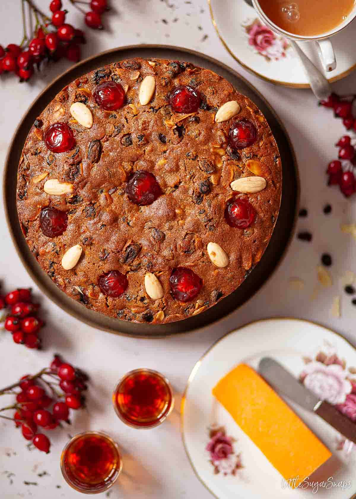 Uncut rum fruit cake topped with glacé cherries and almonds with cheese, rum and tea alongside.