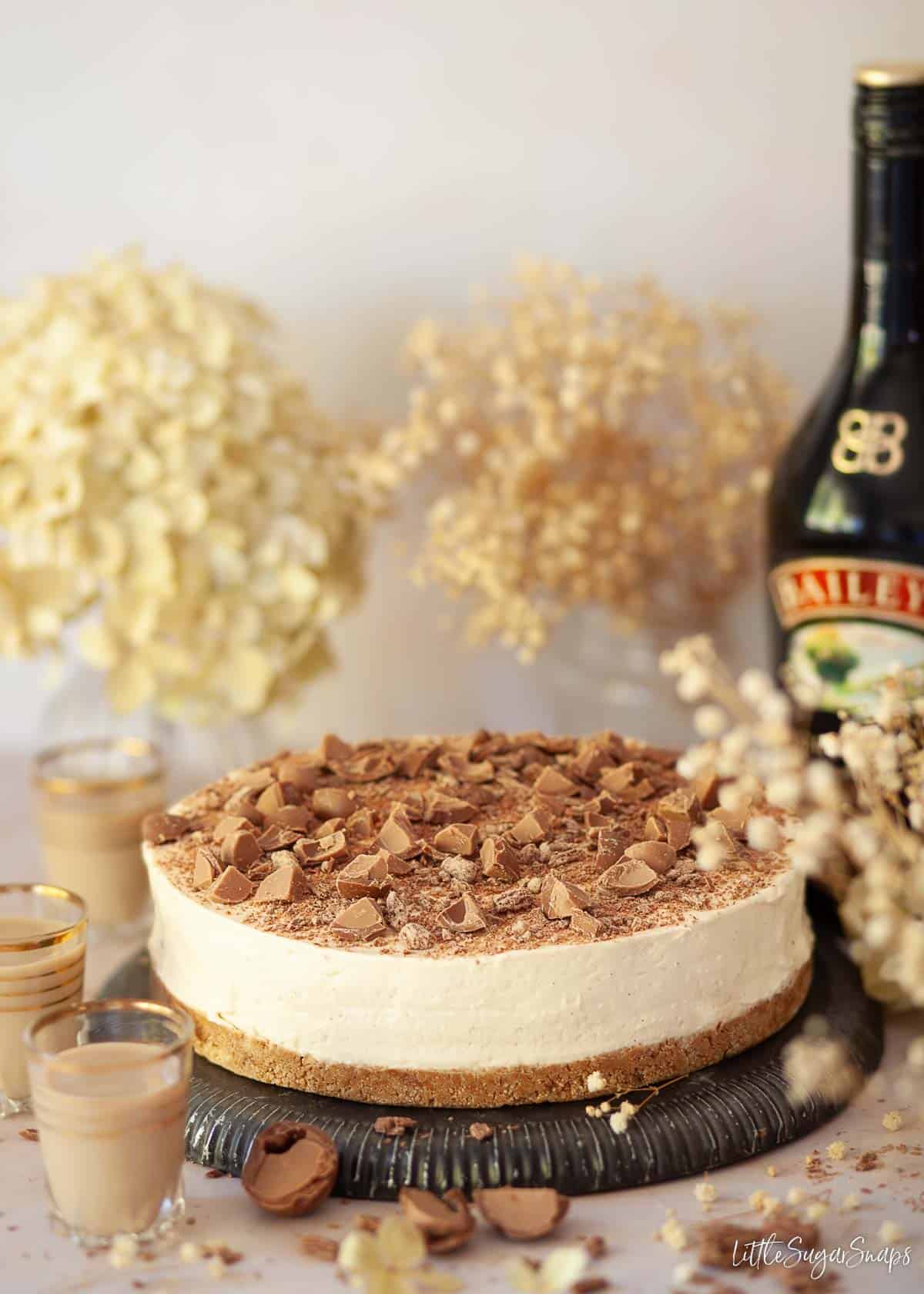 A whole Baileys cheesecake on a plate with a bottle of Baileys Irish cream in the background.