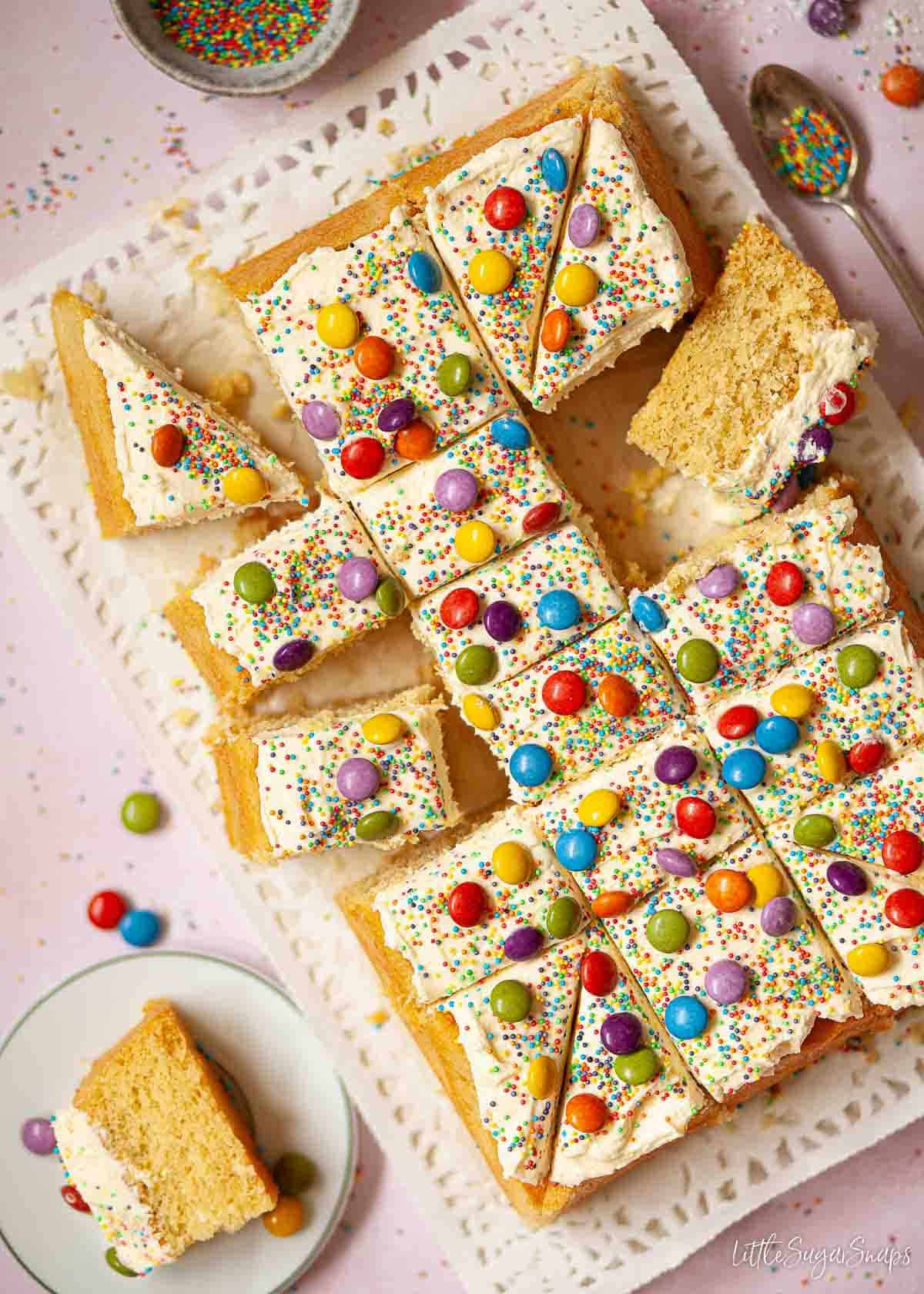 Sliced up vanilla tray bake cake with buttercream, sprinkles and colourful chocolate candies.