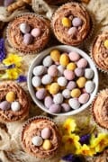 A bowl of mini eggs with spring flowers and small cakes topped with mini eggs around it.