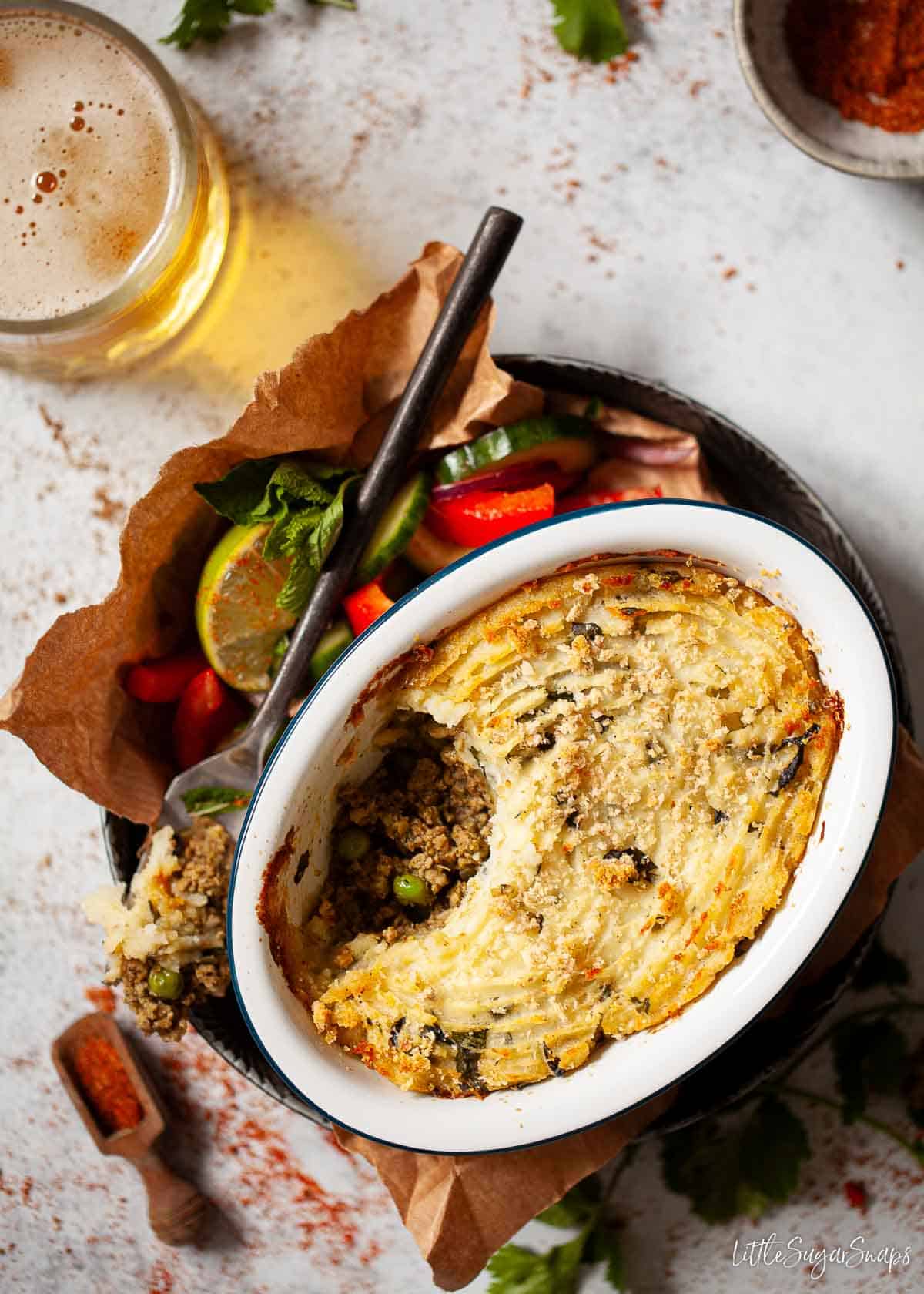 A single serve dish of curried shepherd's pie with side salad and beer.