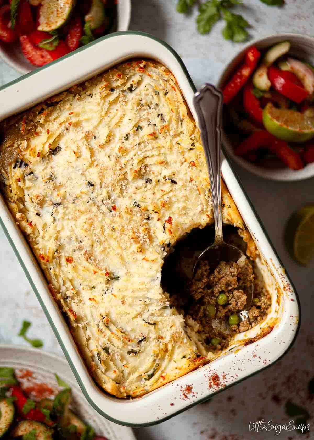 A large minced lamb pie with a portion removed from it and bowls of salad alongside.