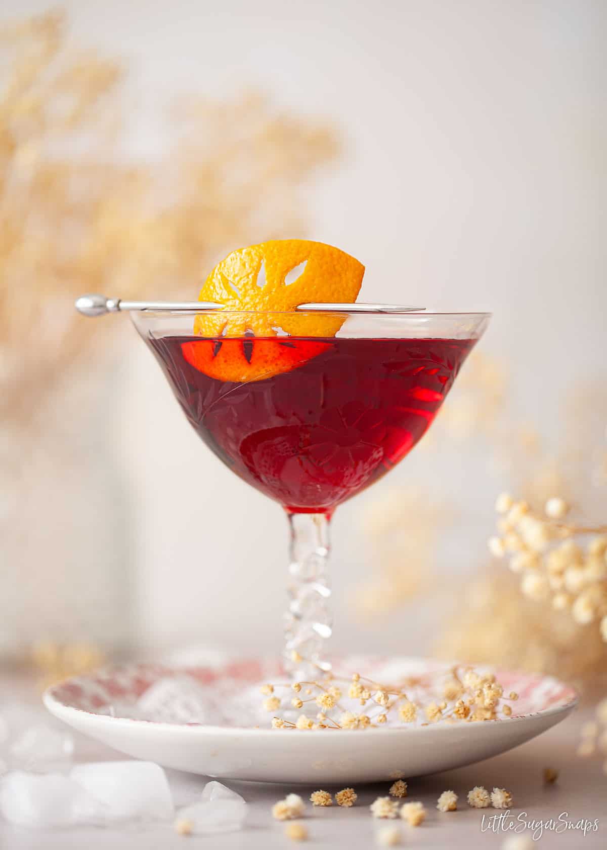 A blackcurrant cocktail in a vintage glass with carved orange zest garnish.