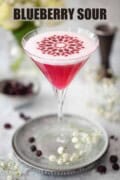 A cocktail garnished with a stencil made from freeze dried blueberry powder with text overlay.