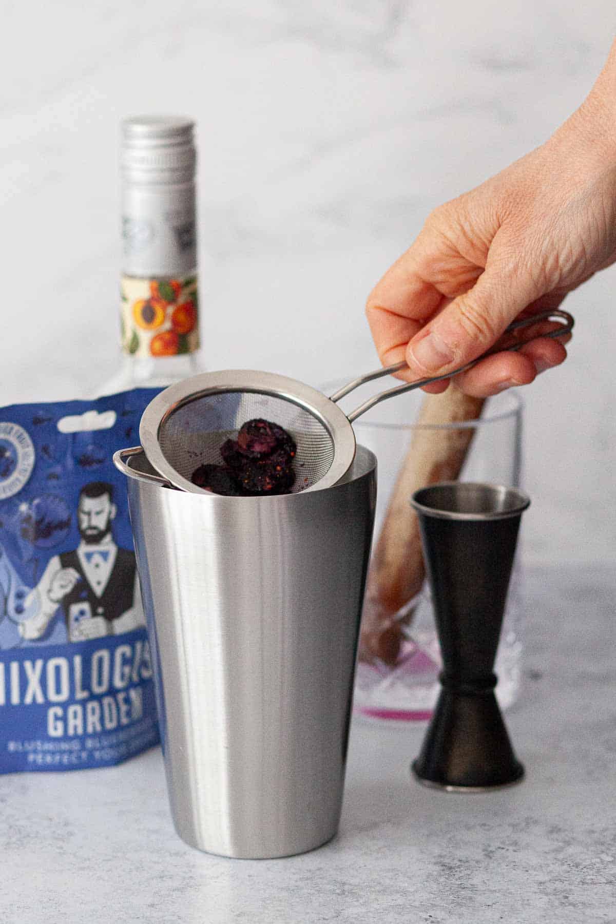 Person holding a sieve over a cocktail shaker with fruit inside of the sieve.