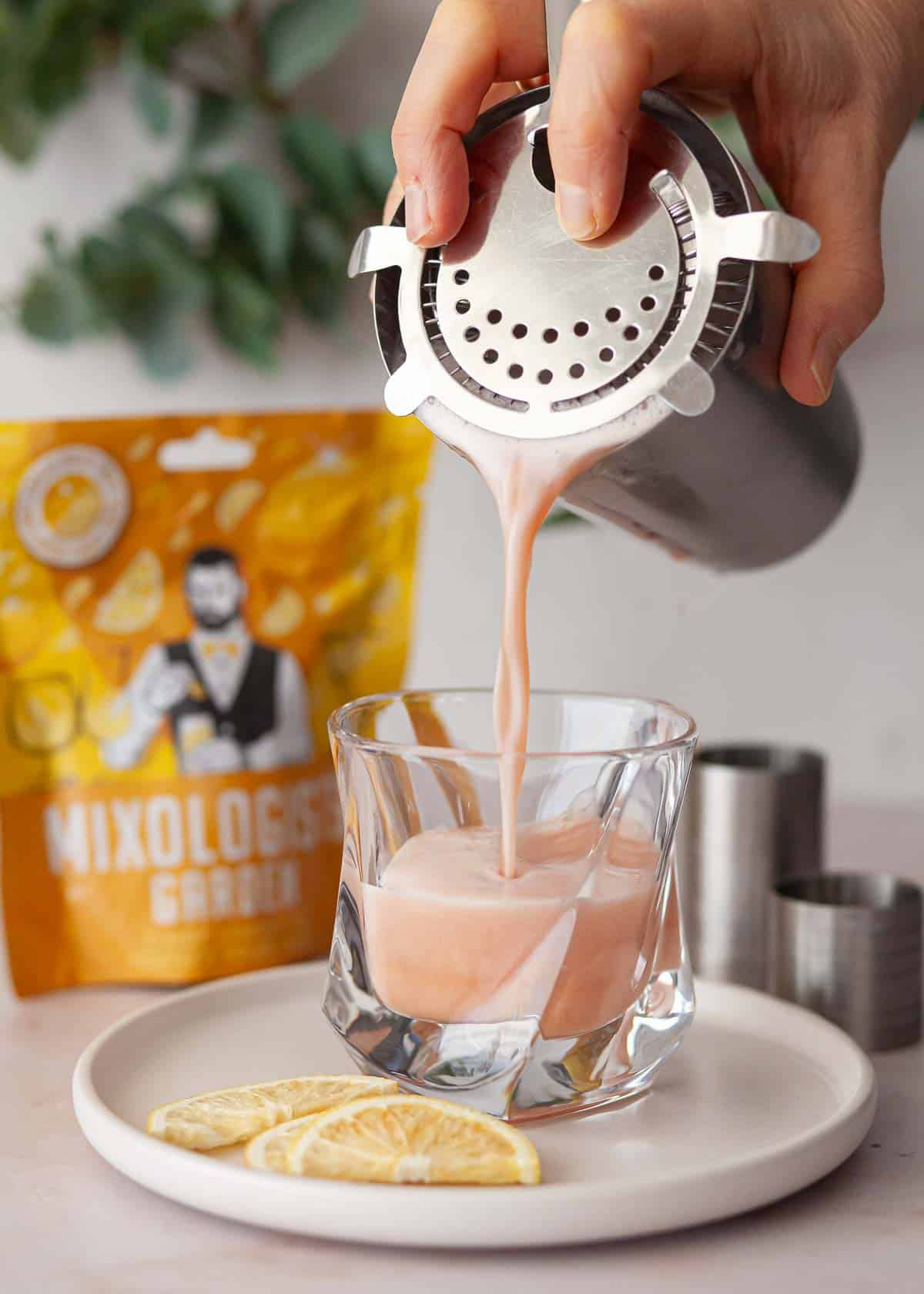 A cocktail being poured through a hawthorne strainer into a tumber.