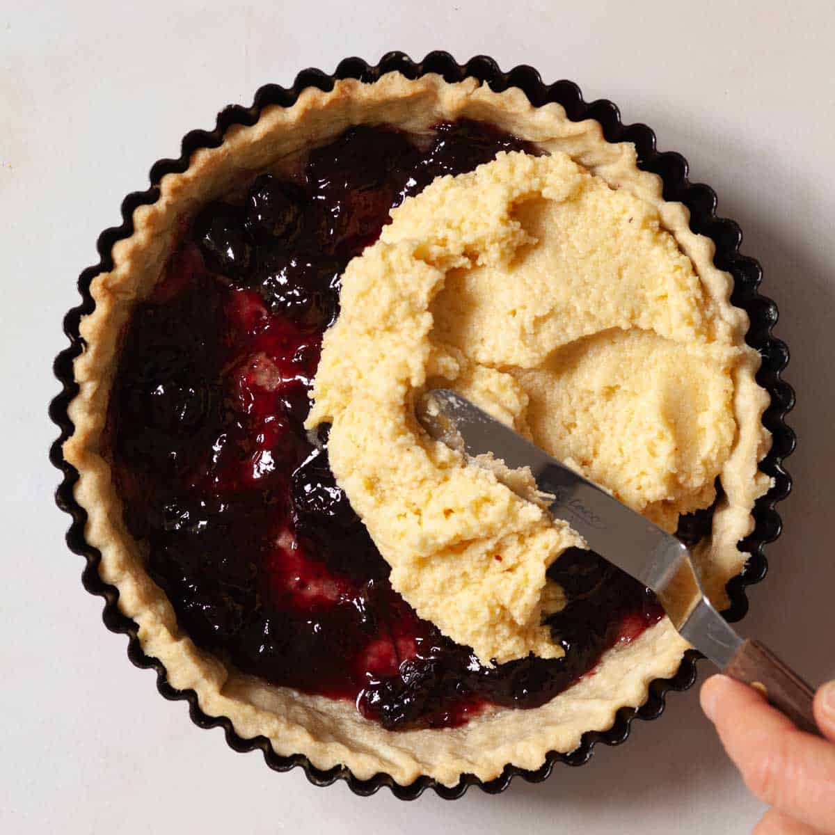 A tart case being filled with jam and frangipane batter.