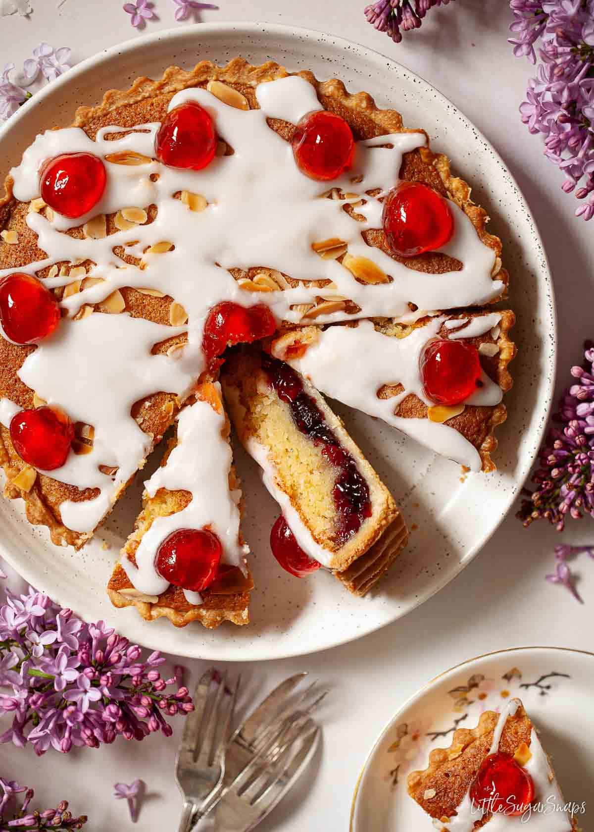 A part-sliced cherry Bakewell tart topped with glacé icing and red glacé cherries.