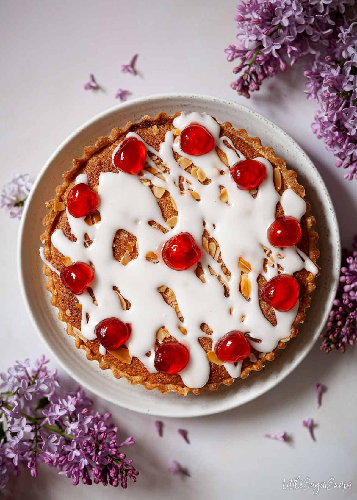 A cherry Bakewell tart topped with glacé icing and red glacé cherries.