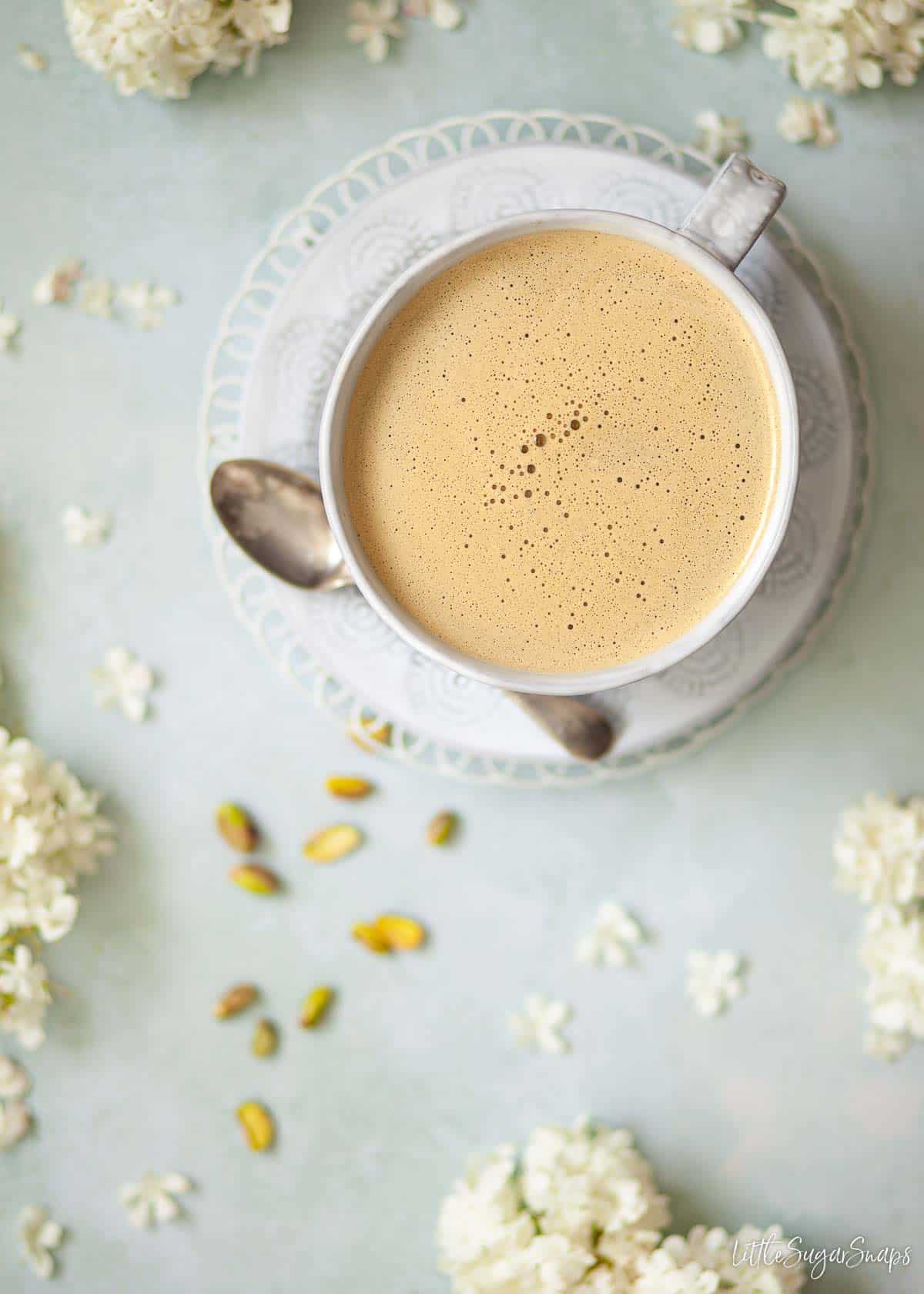 Overhead view of a pistachio coffee in a wide mug with nuts and flowers on the worktop.