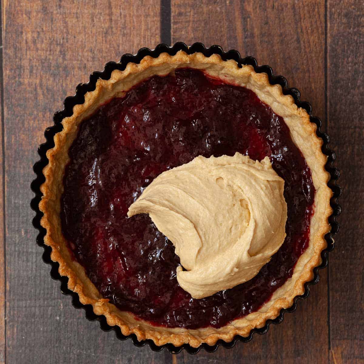Raspberry jam in a baked tart case being topped with sponge cake batter.